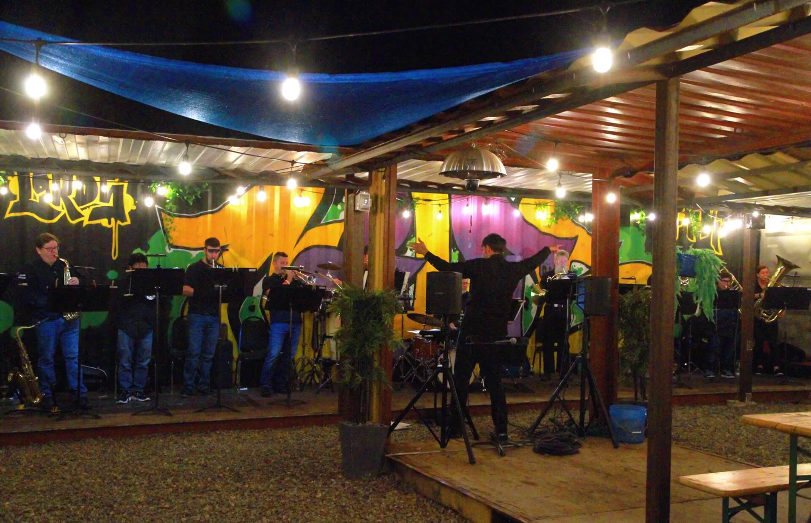 Asheville Symphony Orchestra musicians play brass and saxophones under string lights on an outdoor stage in the River Arts District, with a bright painted wall behind them
