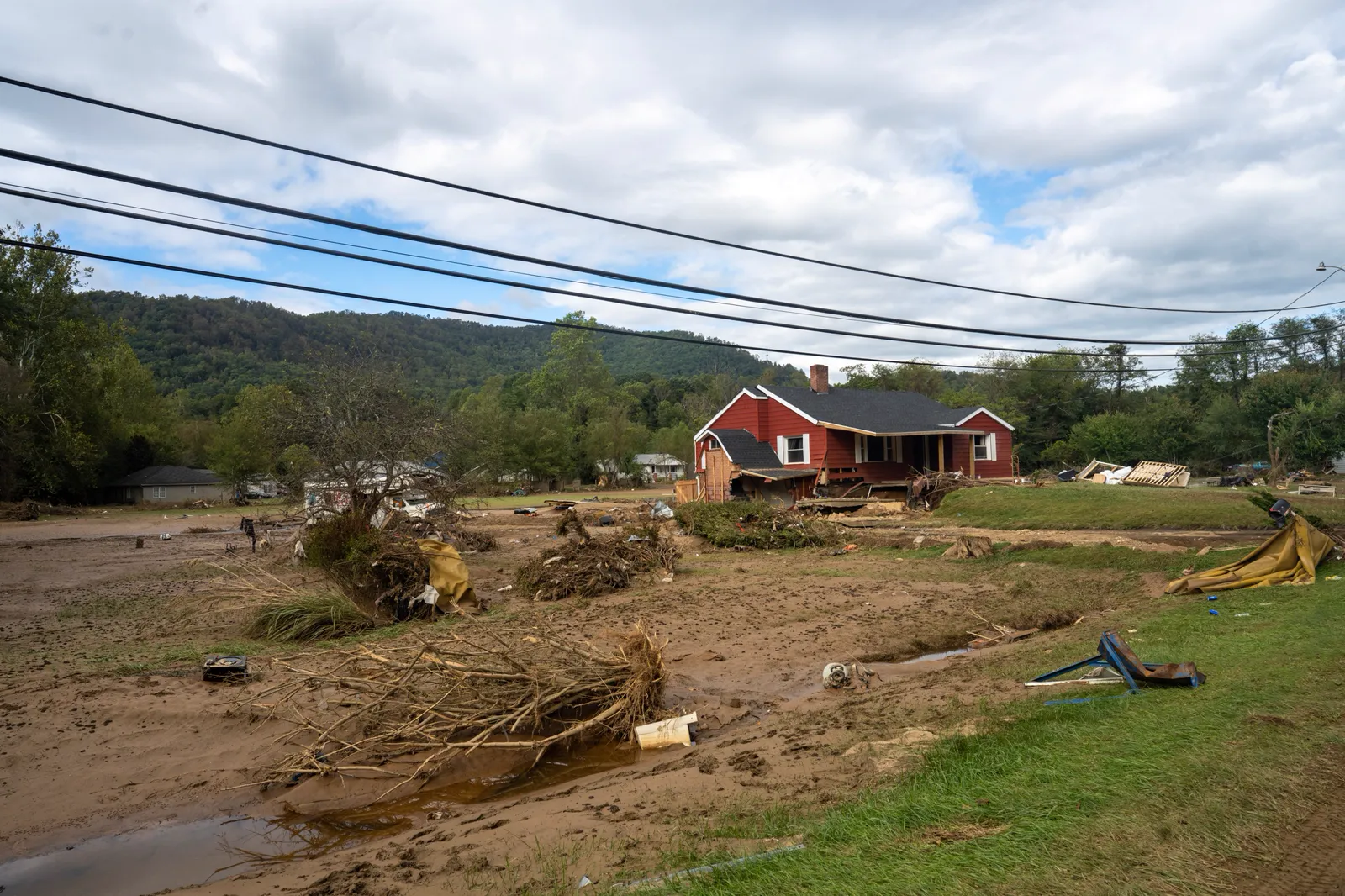 A red house in Swannanoa stands in a mud-covered yard strewn with uprooted brush and debris after Hurricane Helene's flooding, with wooded mountains behind it
