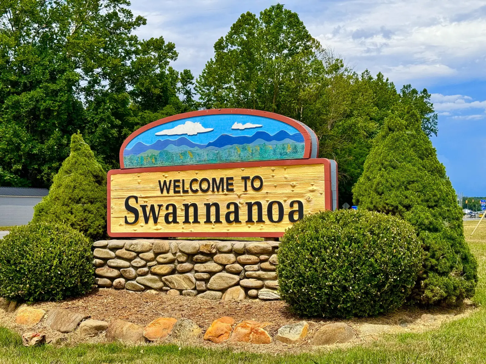 A wooden “Welcome to Swannanoa” sign with a painted mountain scene, set on a stone base among shrubs and trees beside U.S. Highway 70